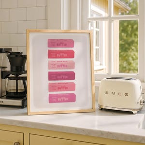 Op de afbeelding: Een ingelijste kunstdruk met zes gestapelde rechthoekige pakjes boter in verschillende tinten roze. Elk pakje is gelabeld met het woord BUTTER en bevat de tekst NET WT 4 OZ (113g). De print zit in een lijst van lichtgekleurd hout.