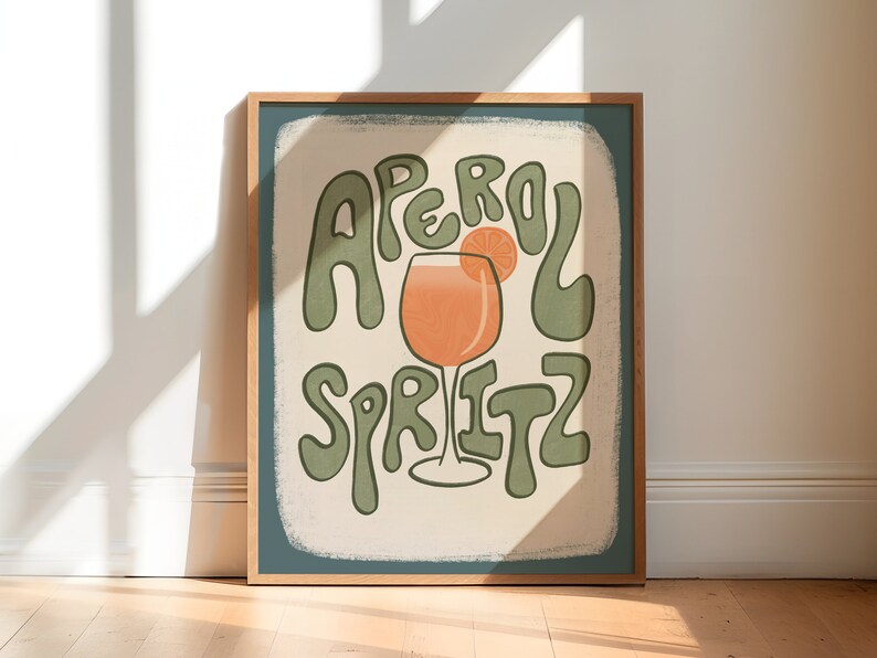 Art Print of an aperol spritz with an orange wedge on the glass. The words "aperol spritz" surround the drink in green. A tan hand drawn rectangle is behind it and the background is a solid color dark teal.