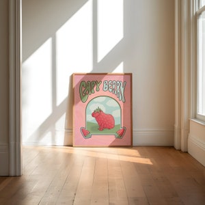 A framed poster with a pink background with green and light blue text that reads 'Capy Berry'. The poster features a cartoon capybara with a strawberry on its head. The capybara is standing on a green patch of grass with a blue sky and white clouds in the background. Two strawberries are on either side of the capybara.