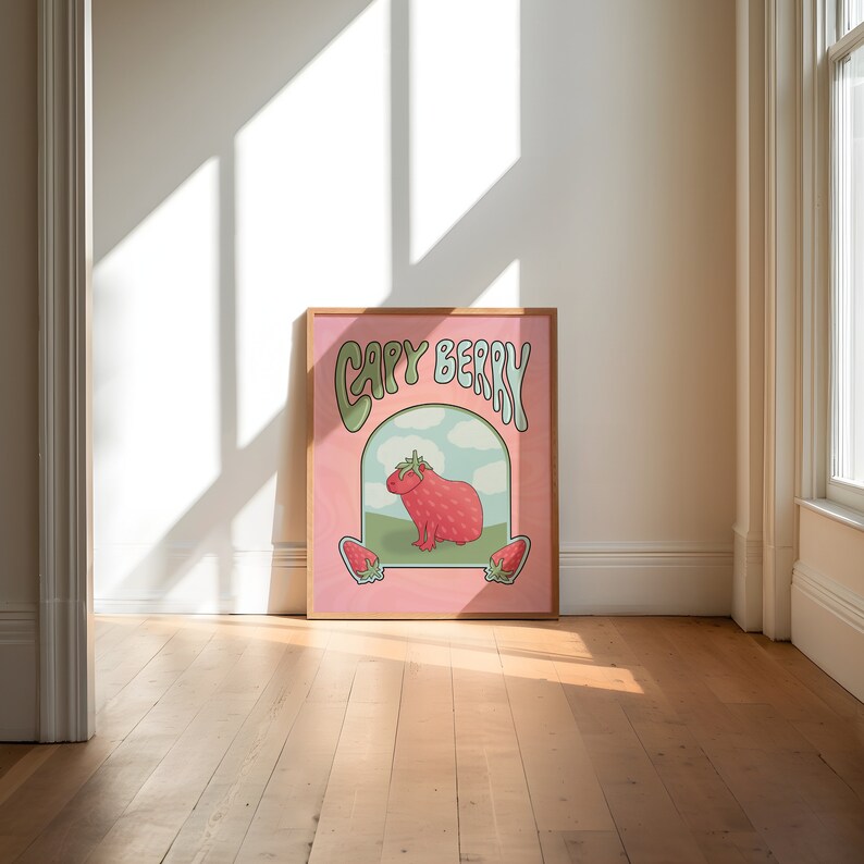 A framed poster with a pink background with green and light blue text that reads 'Capy Berry'. The poster features a cartoon capybara with a strawberry on its head. The capybara is standing on a green patch of grass with a blue sky and white clouds in the background. Two strawberries are on either side of the capybara.