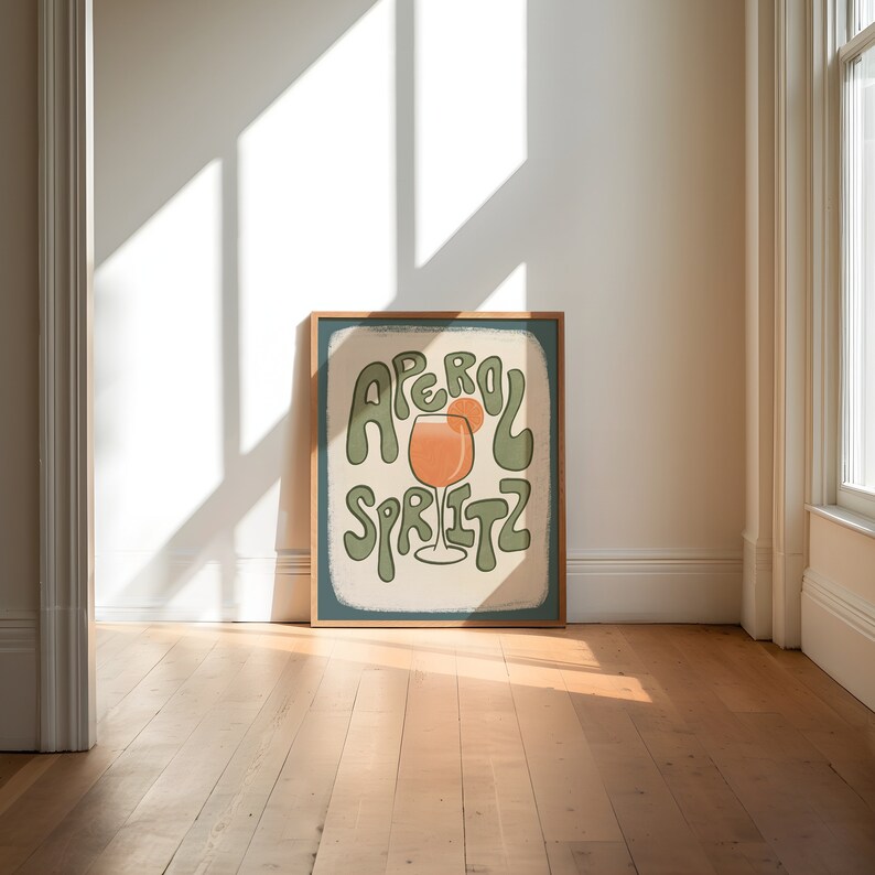 Art Print of an aperol spritz with an orange wedge on the glass. The words "aperol spritz" surround the drink in green. A tan hand drawn rectangle is behind it and the background is a solid color dark teal.