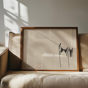 Horizontal art print of a bikini set hanging on a clothesline. The bathing suit is dark grey and in the bottom right of the artwork. The background is tan/beige.