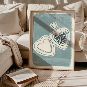 May include: A framed art print featuring a heart-shaped locket design with the text "SAN DIEGO" in a retro font. The locket is open, revealing a blank heart shape. The background is a muted teal color, and the print is in a wooden frame.