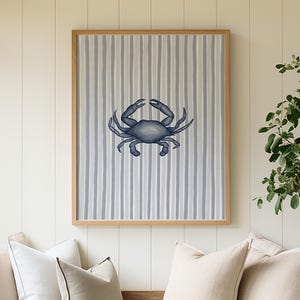 May include: A framed wall art print featuring a centered dark blue crab illustration set against a background of vertical light blue and white stripes. The artwork is enclosed in a simple, light-toned wooden frame, creating a clean visual display.