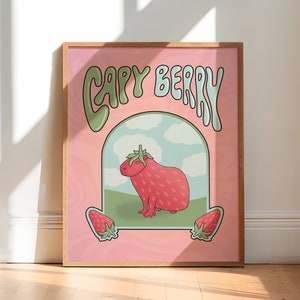 A framed poster with a pink background with green and light blue text that reads 'Capy Berry'. The poster features a cartoon capybara with a strawberry on its head. The capybara is standing on a green patch of grass with a blue sky and white clouds in the background. Two strawberries are on either side of the capybara.