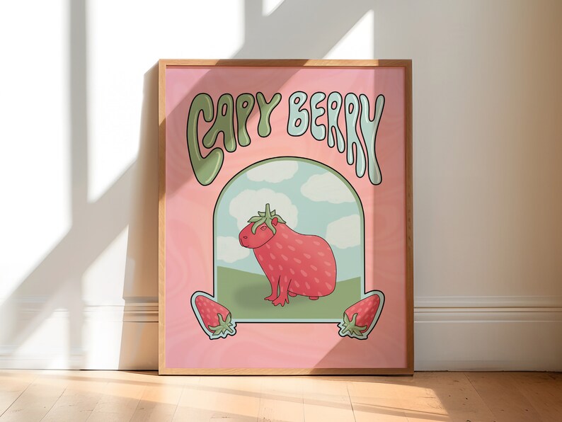 A framed poster with a pink background with green and light blue text that reads 'Capy Berry'. The poster features a cartoon capybara with a strawberry on its head. The capybara is standing on a green patch of grass with a blue sky and white clouds in the background. Two strawberries are on either side of the capybara.