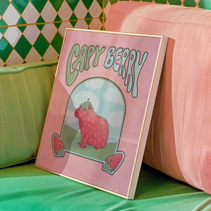 A framed poster with a pink background with green and light blue text that reads 'Capy Berry'. The poster features a cartoon capybara with a strawberry on its head. The capybara is standing on a green patch of grass with a blue sky and white clouds in the background. Two strawberries are on either side of the capybara.