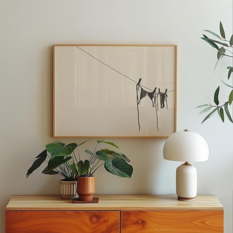 Horizontal art print of a bikini set hanging on a clothesline. The bathing suit is dark grey and in the bottom right of the artwork. The background is tan/beige.