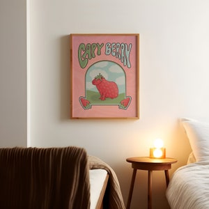 A framed poster with a pink background with green and light blue text that reads 'Capy Berry'. The poster features a cartoon capybara with a strawberry on its head. The capybara is standing on a green patch of grass with a blue sky and white clouds in the background. Two strawberries are on either side of the capybara.