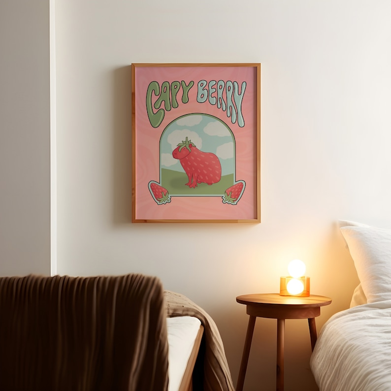 A framed poster with a pink background with green and light blue text that reads 'Capy Berry'. The poster features a cartoon capybara with a strawberry on its head. The capybara is standing on a green patch of grass with a blue sky and white clouds in the background. Two strawberries are on either side of the capybara.