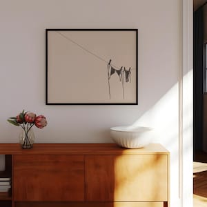 Horizontal art print of a bikini set hanging on a clothesline. The bathing suit is dark grey and in the bottom right of the artwork. The background is tan/beige.