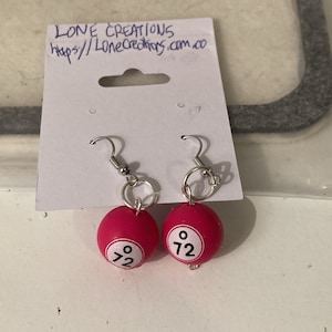 May include: A pair of earrings featuring pink bingo balls with the number 72 printed on them. The earrings are silver and have a hook closure.