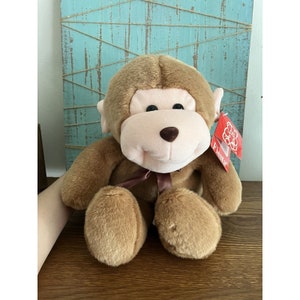 Puede incluir: Un mono de peluche marrón con la cara rosa y los ojos marrones. El mono está sentado sobre una superficie de madera con una etiqueta roja que dice "Fiesta".