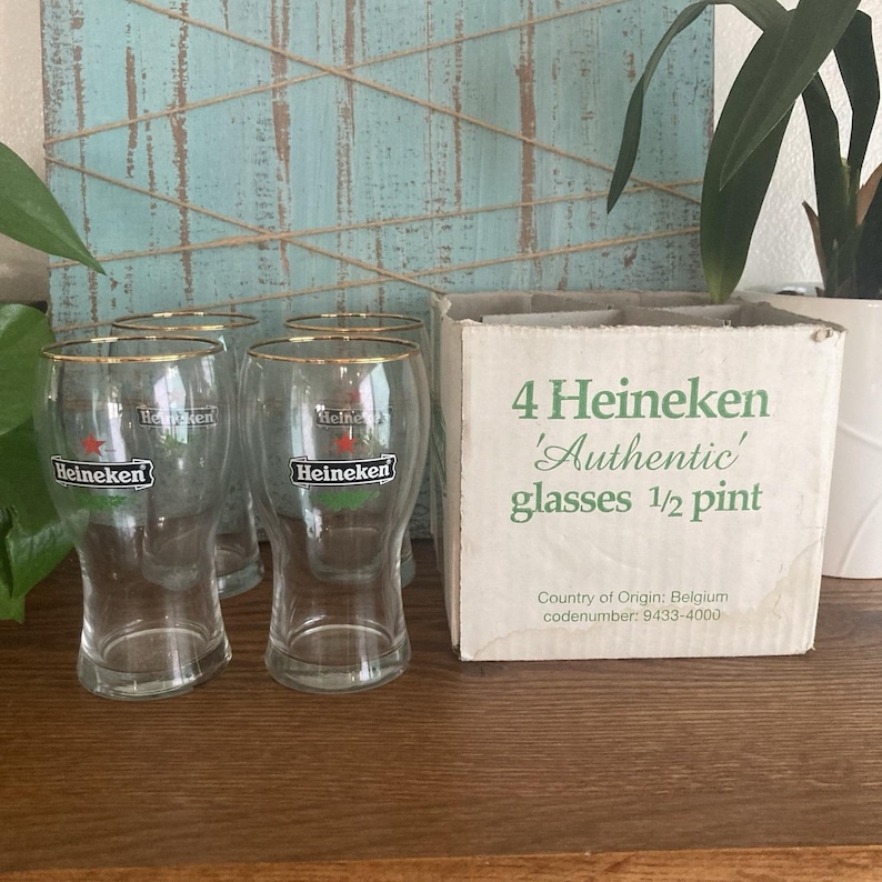 Puede incluir: Cuatro vasos Heineken transparentes con bordes dorados y el logotipo de Heineken, junto a una caja que dice "4 vasos Heineken 'Aut&eacute;nticos' 1/2 pinta". Los vasos est&aacute;n sobre una superficie de madera.