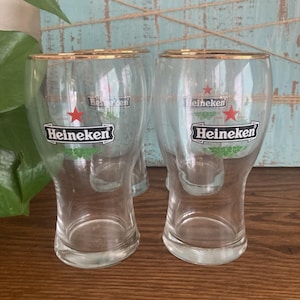 Puede incluir: Vasos de cerveza transparentes con bordes dorados. Cada vaso presenta el logotipo de Heineken con una estrella roja y detalles verdes. Los vasos se exhiben sobre una superficie de madera con un fondo azul claro.