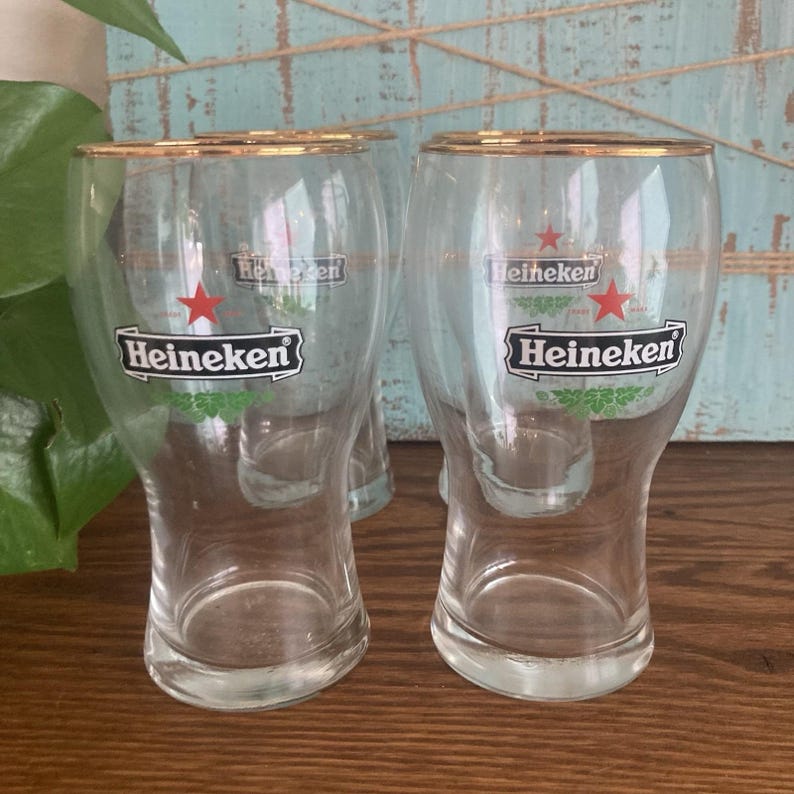 Puede incluir: Vasos de cerveza transparentes con bordes dorados. Cada vaso presenta el logotipo de Heineken con una estrella roja y detalles verdes. Los vasos se exhiben sobre una superficie de madera con un fondo azul claro.