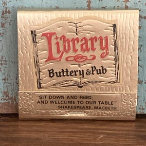 May include: A gold-colored matchbook cover with a textured surface. The front features an illustration of an open book with the red text Library and black text Buttery & Pub. Below, the text reads: SIT DOWN AND FEED, AND WELCOME TO OUR TABLE SHAKESPEARE - MACBETH.