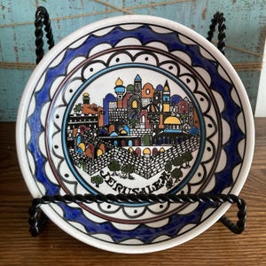 Könnte beinhalten: Ein dekorativer Keramikteller mit einer farbenfrohen Darstellung von Jerusalem. Der Teller hat eine weiße Basis mit einem blauen und braunen Rand. Der Teller wird auf einem schwarzen Metallständer präsentiert. Das Wort "Jerusalem" ist auf dem Teller geschrieben.