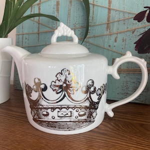 May include: A white ceramic teapot featuring a metallic gold crown graphic on the front. The teapot has a curved handle, a spout, and a removable lid with a decorative finial handle. The design is centered on the body of the white teapot.