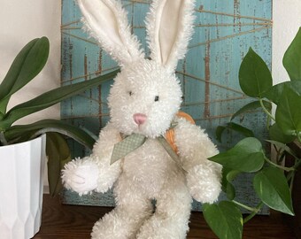 May include: A plush toy rabbit with cream-colored, textured fur, long upright ears, and a pink nose. The rabbit wears a light green and white checkered ribbon tied around its neck and carries a small orange backpack on its back.