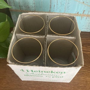 Puede incluir: Un juego de cuatro vasos transparentes con bordes dorados, presentados en una caja de cart&oacute;n. La caja lleva la inscripci&oacute;n "4 Heineken Authentic Glasses 1/2 Pint" en texto verde. Los vasos son para cerveza.