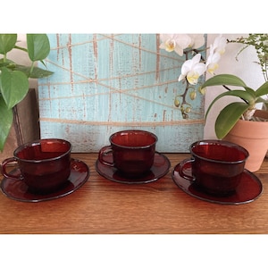 May include: Three red glass teacups and saucers. The cups are a deep red color and have a rounded shape. The saucers are a lighter shade of red and have a scalloped edge.