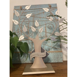 May include: A silver metal tree sculpture with leaves that have words on them, such as "love", "joy", "life", and "forever close in heart". The tree is on a wooden base.