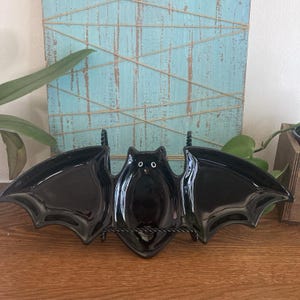 May include: A glossy black ceramic serving dish shaped like a bat with outstretched wings. The central body features two small white circular eyes. The dish is displayed on a black metal stand, showcasing a wide, symmetrical design.