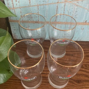 Puede incluir: Cuatro vasos de cerveza transparentes con bordes dorados. Cada vaso tiene el logotipo de Heineken impreso en la parte delantera. Los vasos est&aacute;n sobre una superficie de madera con un fondo azul y blanco.