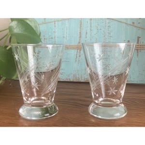May include: Two clear, etched glass tumblers with a starburst design. The glasses have a flared shape and a rounded base. The etched design features star shapes and curved lines. The glasses are displayed on a wooden surface.