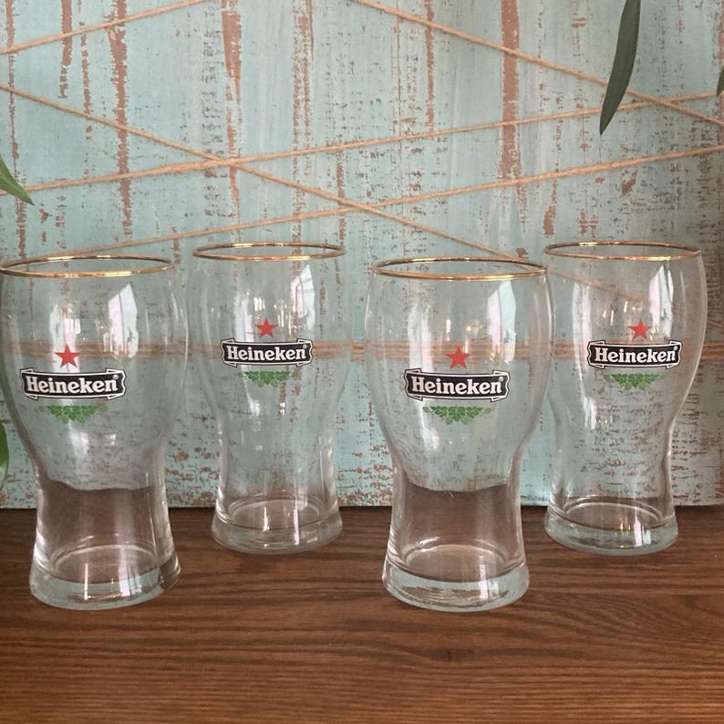 Puede incluir: Cuatro vasos de cerveza transparentes con bordes dorados. Cada vaso presenta el logotipo de Heineken con una estrella roja y detalles verdes. Los vasos est&aacute;n sobre una superficie de madera con un fondo azul claro.