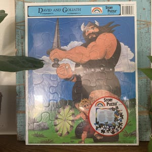 Op de afbeelding: Een David en Goliath Story Puzzle met een cartoonillustratie van een grote, gespierde reus met een zwaard en een kleinere figuur eronder. De puzzel bevat een ronde afbeelding en tekst bovenaan, verpakt in doorzichtig plastic folie.