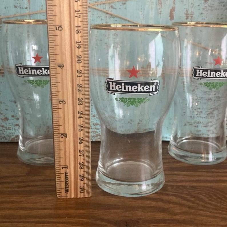 Puede incluir: Tres vasos de cerveza transparentes con bordes dorados, cada uno con el logotipo de Heineken con una estrella roja y detalles verdes. Una regla de madera se coloca junto a los vasos. Los vasos miden aproximadamente 15 cm de alto.
