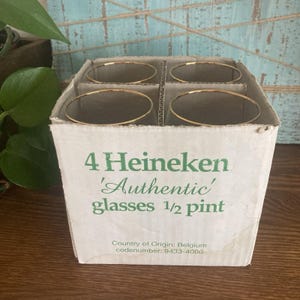 Puede incluir: Una caja de cart&oacute;n con cuatro vasos Heineken 'Authentic' con bordes dorados. La caja tiene texto verde que dice "4 Heineken 'Authentic' glasses 1/2 pint". Cada vaso tiene aproximadamente 284 ml.
