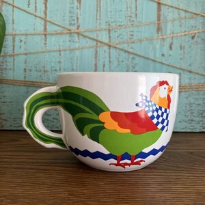 Peut inclure: Une tasse en céramique blanche avec un motif de coq. Le coq est vert, rouge, orange et jaune avec une écharpe à carreaux bleus et blancs. La tasse a un fond blanc avec des lignes ondulées bleues et blanches autour du bas.
