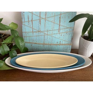 以下が含まれることがあります： 3つの楕円形のセラミック製サービングプラッターのスタック。一番下のプラッターは白、真ん中は青、一番上は薄い黄色です。プラッターは木製の表面にあり、背景にはターコイズブルーの木製パネルがあります。