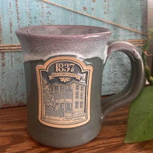May include: A ceramic mug with a two-tone gray and lavender glaze. The front features a raised, tan-colored emblem depicting a multi-story building with the text 1837 BED & BREAKFAST and Charleston, South Carolina printed below the image.