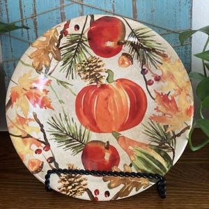 May include: A round ceramic plate featuring a seasonal autumn motif with a central orange pumpkin, red apples, brown pinecones, acorns, green pine needles, and yellow and orange maple leaves on a cream-colored background, displayed on a black stand.