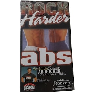 May include: A fitness video box with the title "ROCK Harder" in red and white lettering. The word "abs" is prominently displayed. The box features images of toned abs and a man in a black shirt. The text reads "the ultimate 5 minute AB ROCKER workout video".