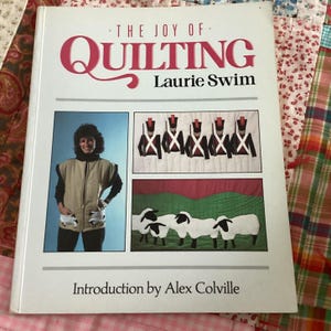May include: A book titled "The Joy of Quilting" by Laurie Swim. The cover features a woman in a tan vest, and images of quilts with soldiers and sheep. Introduction by Alex Colville.