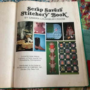 Puede incluir: Libro abierto titulado "Scrap Savers' Stitchery Book" de Sandra Lounsbury Foose. El libro presenta varios proyectos de costura, incluyendo un hipopótamo rosa, un elefante azul, un granero rojo y una pila de monos. Publicado por Countryside Press.