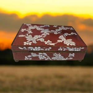 May include: A rectangular, red jewelry box with a floral design in white. The box has a hinged lid and small feet. The floral pattern covers the top and sides of the box. The background is a blurred image of a sunset.