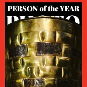 May include: A gold-colored trophy with a textured surface. The trophy is shaped like a tall, cylindrical cup with a wide base. The text "PERSON of the YEAR" is visible at the top of the image.
