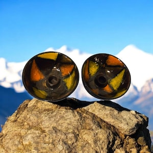 May include: Two black ceramic candle holders with a geometric design in yellow and orange. The holders are sitting on a rock with a mountain background.