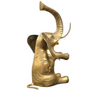 Pode incluir: Uma escultura de elefante de latão dourado com a tromba levantada no ar. O elefante está sentado sobre suas patas traseiras com as patas dianteiras levantadas. A escultura é detalhada com linhas e rugas texturizadas.