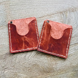 May include: Two brown leather card holders with a stitched edge and a cut-out for a card. The holders are on a wood surface.