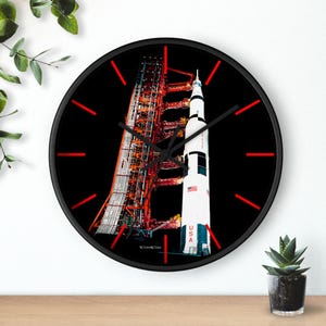 Könnte beinhalten: Schwarze runde Wanduhr mit einem detaillierten Bild der Saturn V-Rakete auf ihrer Startrampe. Die Uhr hat rote Stundenmarkierungen und schwarze Zeiger. Die Rakete ist weiß mit schwarzen Streifen und der US-Flagge.