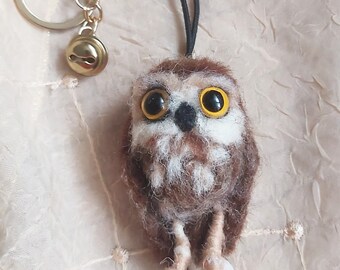 May include: A handcrafted owl keychain with a gold-colored bell and keyring. The owl is made of brown and white felted wool, featuring large, expressive eyes with yellow irises. The owl's feet are visible, and it hangs from a black cord.