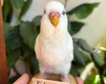 Puede incluir: Figurita de pájaro de fieltro blanco hecha a mano, con pico amarillo y detalles rosas. El pájaro está posado sobre una pequeña base de madera con el nombre "Fatmanur" en un pequeño letrero. Fondo verde.