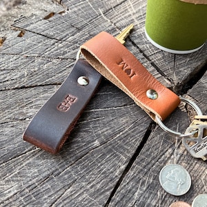 May include: Two leather keychains, one dark brown and one tan, each with initials stamped on them. The keychains are attached to metal key rings. Keys and coins are also visible, all resting on a weathered wooden surface.